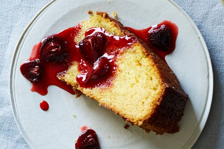 A slice of vanilla cake sits on a white plate. Roasted strawberries, leaking red juice, have been artfully strewn across the slice.